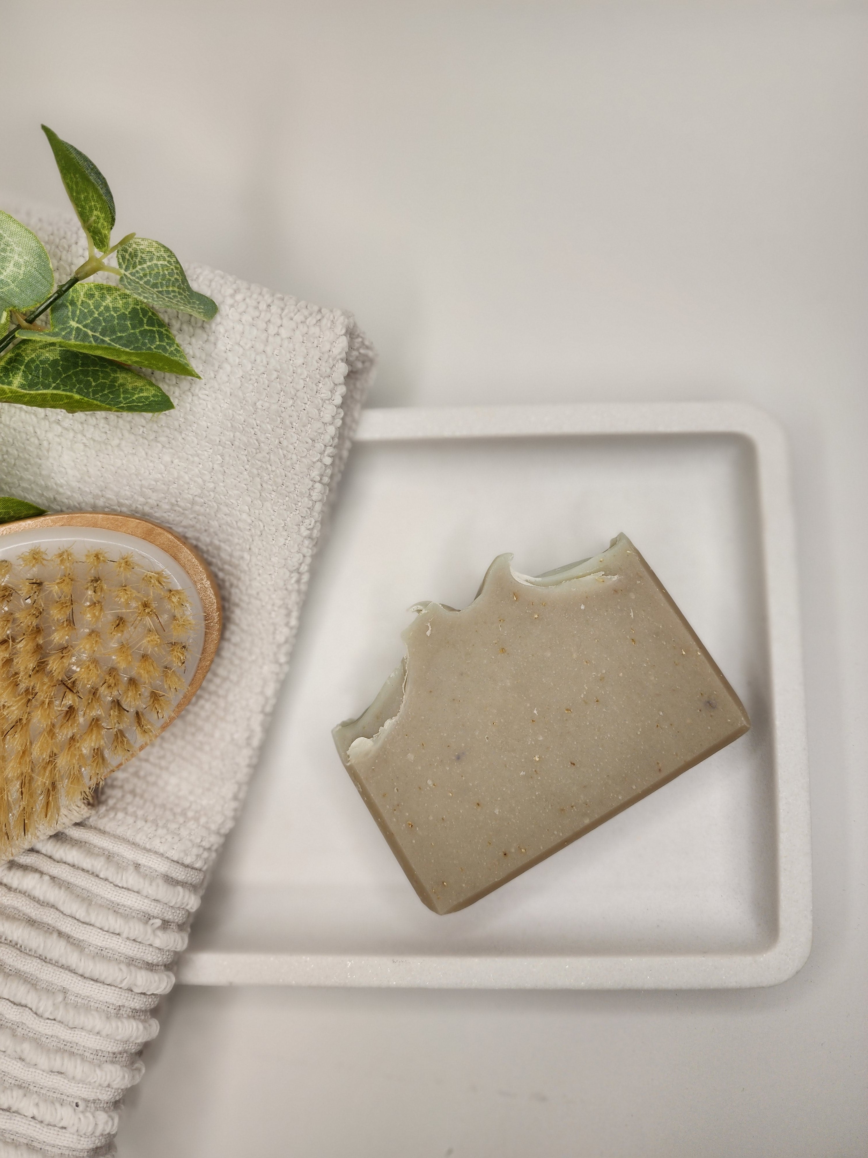 A grey bar of Lavender + Oatmeal handmade soap on a white platter next to a light grey towel and brush